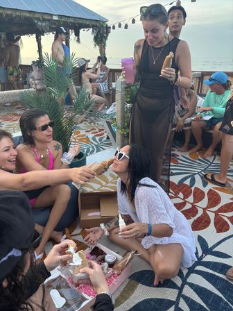 Group of friends at a beachfront tiki-style bar laughing and sharing novelty pastries, woman in heart-shaped sunglasses kneeling as others hand a pastry, colorful patterned rug and ocean sunset in the background