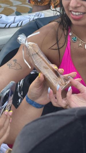 Poolside close-up of a person in a pink bikini and shell charm necklace holding a clear-wrapped brown pastry or cigar-shaped snack while another hand photographs it on a phone, towels and lounge chair in the background.