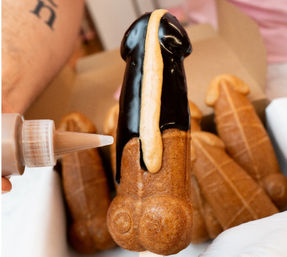 Playful penis-shaped choux pastry on a stick dipped in glossy dark chocolate with a caramel drizzle, shown in a pastry box with other novelty pastries.