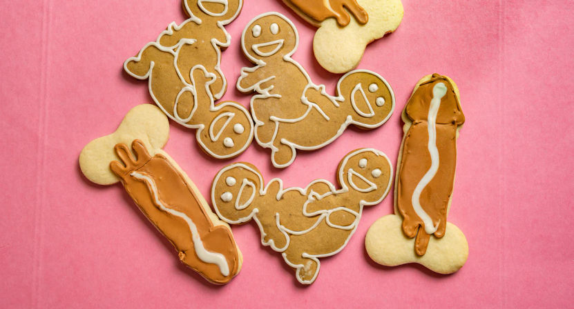 Novelty adult-themed sugar cookies: smiling gingerbread figures and chocolate‑iced phallic cookies with white icing, arranged on a pink background