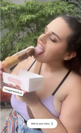 Young woman in a lavender tank top enjoying a chocolate-dipped ice pop outdoors, licking the treat while holding a white takeout box against a leafy background.