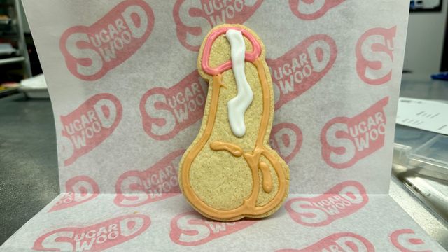 Humorous penis-shaped sugar cookie decorated with pink, orange, and white icing, displayed upright on bakery parchment paper over a stainless countertop.