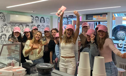 Group of women wearing pink caps celebrating inside a bright dessert shop, smiling and holding sweets behind the counter with stacked paper cups and a pastry display.