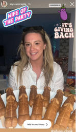 Smiling woman at a bachelorette-style party holding a plate of novelty penis-shaped pastries with Instagram stickers reading "WIFE OF THE PARTY" and "IT'S GIVING BACH" and snacks on a kitchen counter in the background.