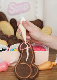 Hand piping white icing onto a chocolate, adult-themed cookie shaped like a penis, with pink and brown icing details, colorful sprinkles and icing bags nearby.