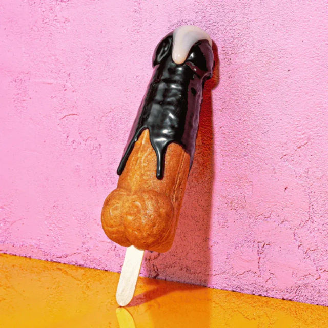 Novelty phallic popsicle dessert on a wooden stick, cake-like base dipped in dark chocolate with a white drip, photographed against a pink textured wall and orange surface