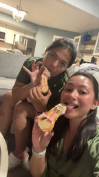 Two friends taking a selfie on a couch in an open-plan living room and kitchen, playfully licking decorated novelty cookies iced with candy eyes and pink details.