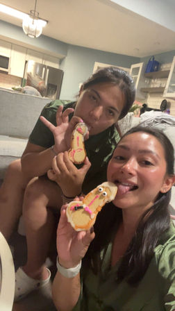 Two friends taking a selfie on a couch in an open-plan living room and kitchen, playfully licking decorated novelty cookies iced with candy eyes and pink details.