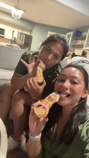 Two friends taking a selfie on a couch in an open-plan living room and kitchen, playfully licking decorated novelty cookies iced with candy eyes and pink details.