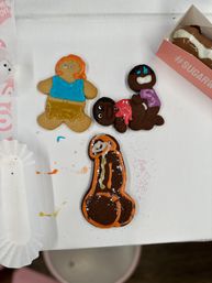 Assorted decorated cookies on a white box — two iced gingerbread people and a chocolate cookie outlined in orange in a playful adult-themed phallic shape, with colorful icing and sprinkles.