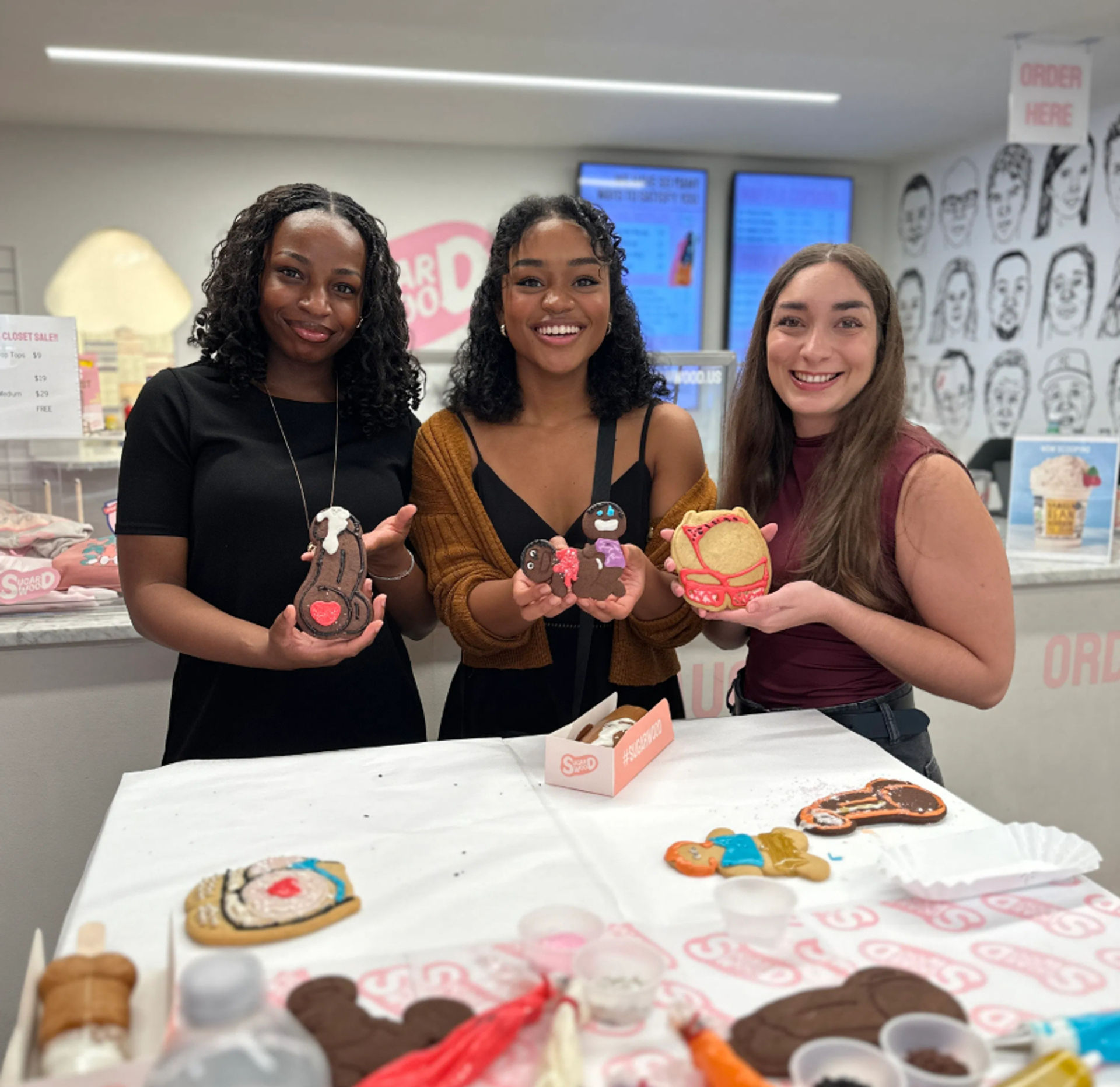Three smiling people at a cookie-decorating counter in a bright dessert shop holding colorful decorated cookies, with icing cups and extra cookies on the table and menu screens and illustrated wall art in the background.