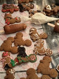 Festive gingerbread cookie decorating scene on a kitchen counter: assorted homemade gingerbread people on parchment with colorful royal icing details and a piping bag.