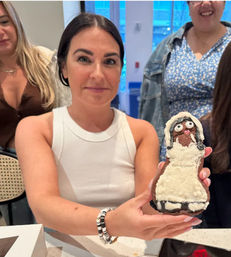 Person holding a decorated gingerbread bride cookie with white icing dress and candy eyes, friends gathered in a bright indoor space.