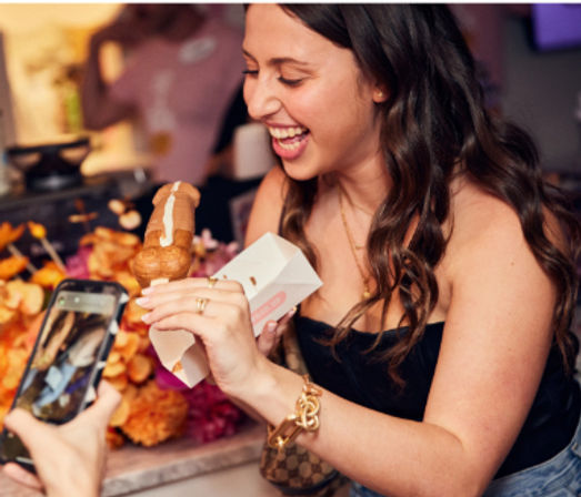 Laughing woman at a dessert counter enjoying a cream-filled pastry on a stick while a friend photographs it with a smartphone, colorful floral display in the background.