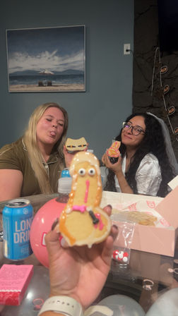 Two friends at an indoor bachelorette-style party smiling and holding playful decorated novelty cookies, a googly-eyed cookie is in the foreground and a beach painting hangs on the wall behind them.