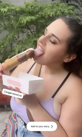 Person in a lavender top licking a chocolate-dipped ice cream bar outdoors while holding a dessert box, playful summer snack