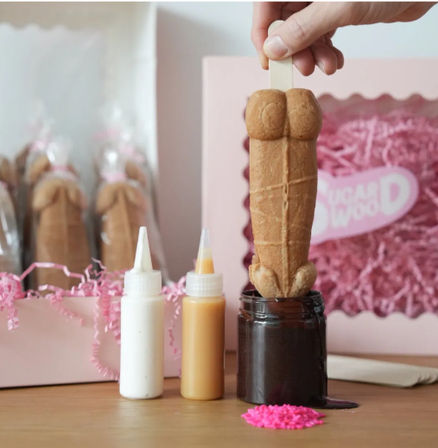 Hand dipping a novelty waffle dessert pop into a jar of chocolate sauce, with white and caramel drizzle bottles, pink sprinkles and a pink gift box in the background.