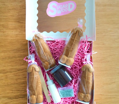 Bachelorette party favor box on wooden table with four individually wrapped novelty penis-shaped cookies on pink shredded paper, plus small icing bottle, chocolate jar, sprinkles packet and wooden applicator.