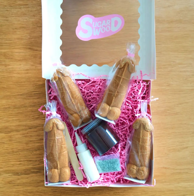 Bachelorette party favor box on wooden table with four individually wrapped novelty penis-shaped cookies on pink shredded paper, plus small icing bottle, chocolate jar, sprinkles packet and wooden applicator.