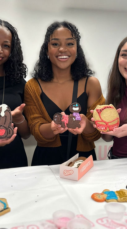 Three smiling friends at an indoor cookie-decorating workshop, each holding colorful decorated sugar cookies over a table with icing supplies