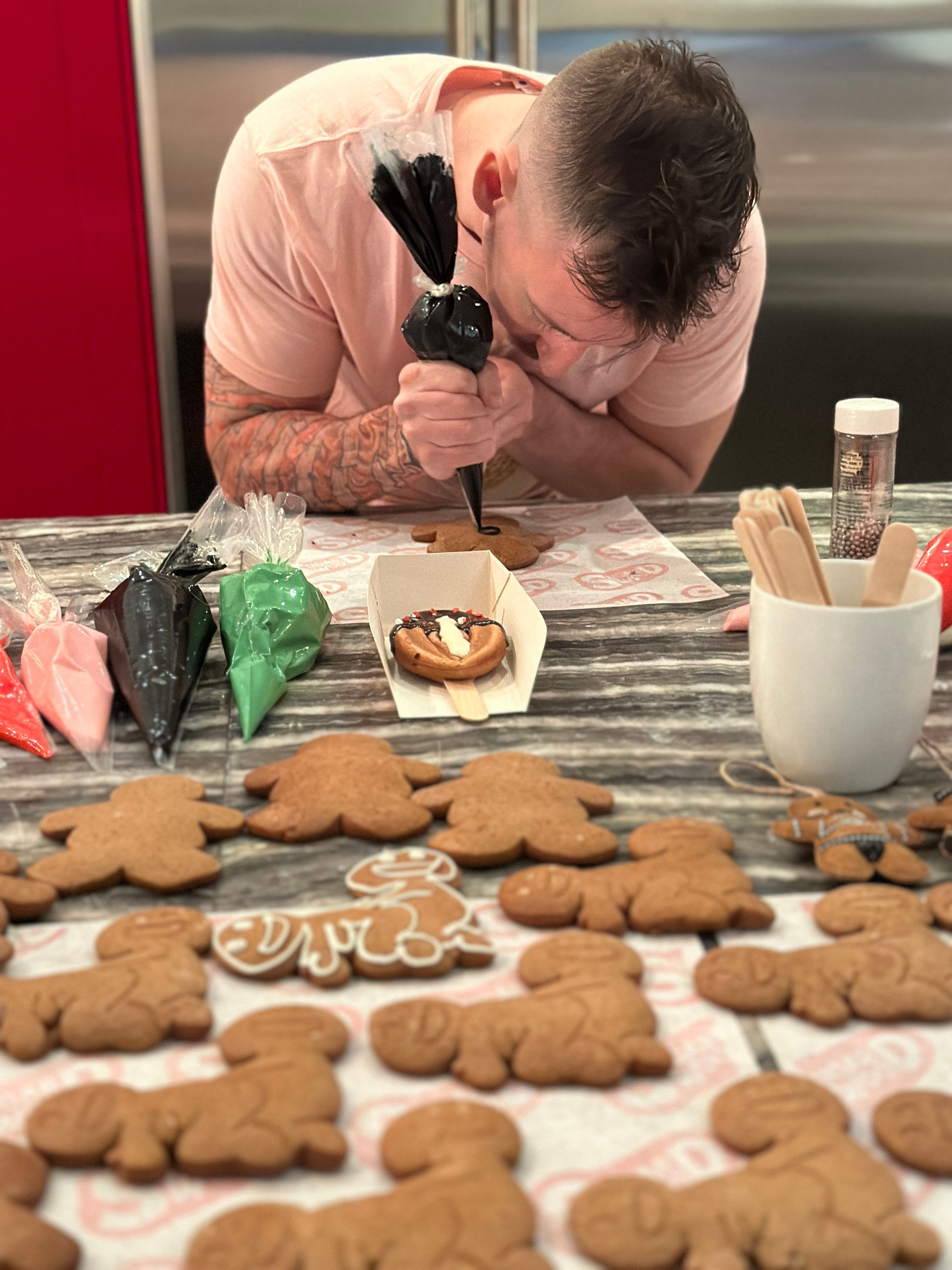 Person at a home kitchen counter piping black icing onto a gingerbread cookie, surrounded by uniced gingerbread men, colorful icing bags, sprinkles and wooden utensils - holiday cookie decorating.