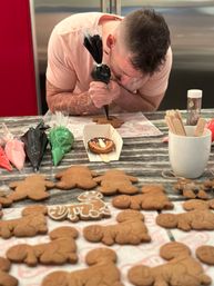 Person at a home kitchen counter piping black icing onto a gingerbread cookie, surrounded by uniced gingerbread men, colorful icing bags, sprinkles and wooden utensils - holiday cookie decorating.