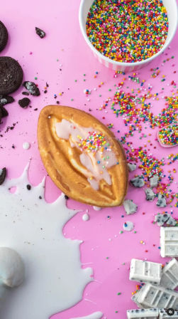 Playful dessert flatlay: boat-shaped waffle drizzled with white glaze and rainbow nonpareil sprinkles on a bright pink background, surrounded by spilled milk, crumbled chocolate sandwich cookies, white chocolate chunks, and a bowl of multicolored sprinkles.