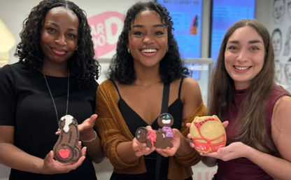 Three smiling women indoors holding colorful decorated novelty cookies—character, heart and mask-shaped treats from a cookie-decorating activity.