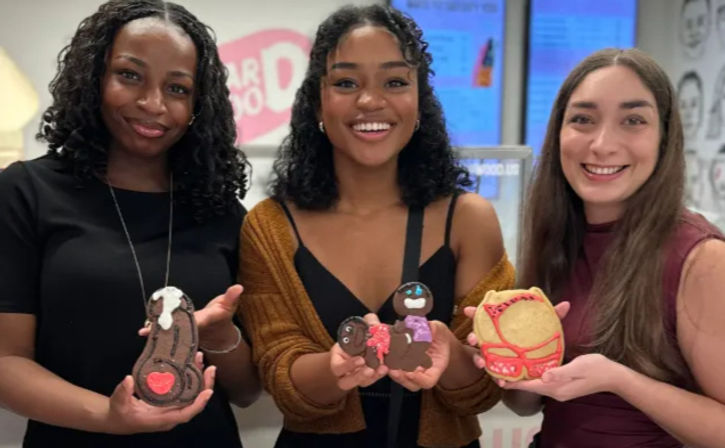 Three smiling women indoors holding colorful decorated novelty cookies—character, heart and mask-shaped treats from a cookie-decorating activity.