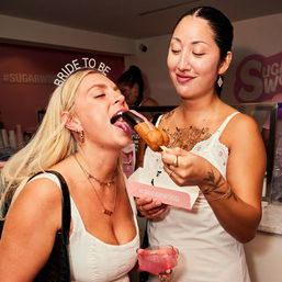Two women at a bachelorette-style party in a dessert shop — one wearing a 'BRIDE TO BE' headband playfully bites a chocolate-dipped pastry on a stick held by her friend, with a pink party cup and takeout box visible.
