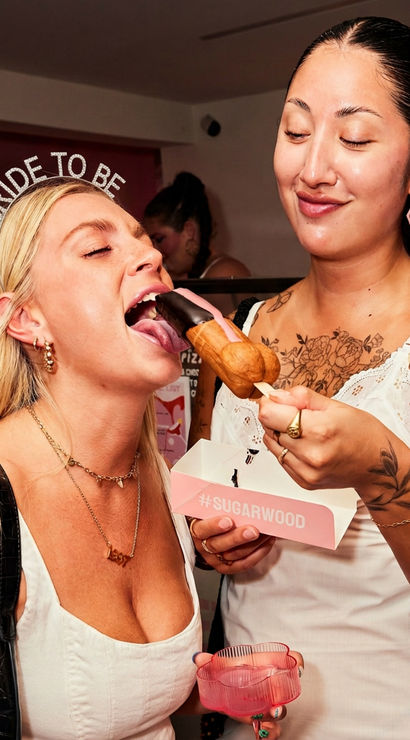 Two women at a bachelorette-style party in a dessert shop — one wearing a 'BRIDE TO BE' headband playfully bites a chocolate-dipped pastry on a stick held by her friend, with a pink party cup and takeout box visible.