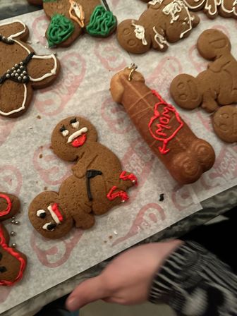 Close-up of holiday gingerbread cookies on parchment at a cookie-decorating table — iced gingerbread people, a novelty adult-shaped cookie with red icing heart, and a hand reaching in.