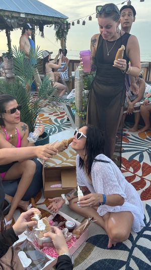 Group of friends at an oceanfront beach bar deck, laughing on a colorful patterned rug as one woman in heart-shaped sunglasses kneels while others feed her cream-filled pastry cones, tropical plants, string lights and sea view in the background.
