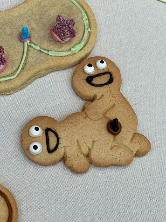 Close-up of a playful gingerbread cookie shaped like two joined figures with candy eyes and black icing smiles on a white tablecloth, next to a partially visible decorated cookie with green icing and pink star candy