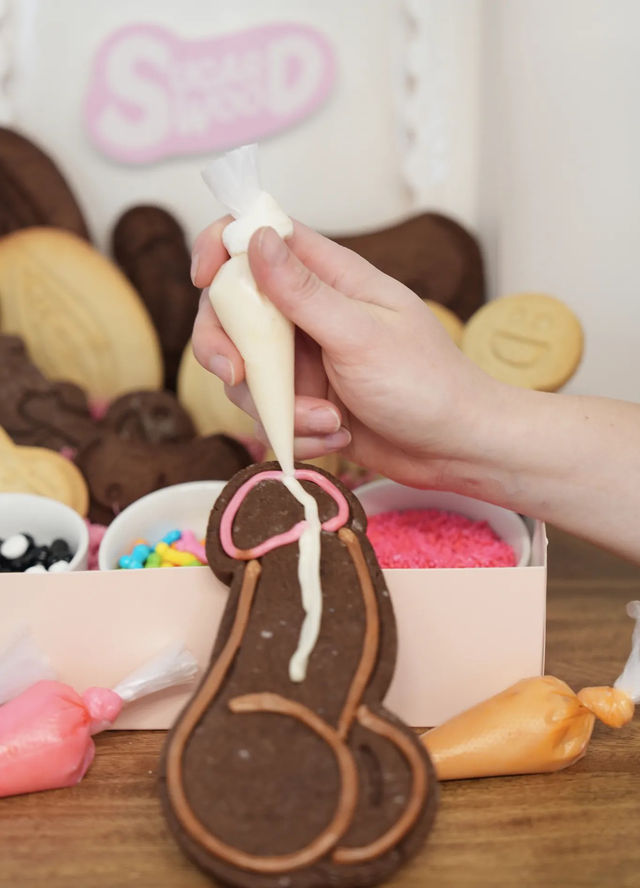 Hand piping white icing onto a chocolate novelty penis-shaped cookie in a DIY cookie-decorating kit with colorful sprinkles and icing bags on a wooden table