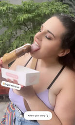 Close-up of a young woman in a lavender top enjoying a chocolate-dipped ice cream bar outdoors, holding a white dessert box against a leafy background.