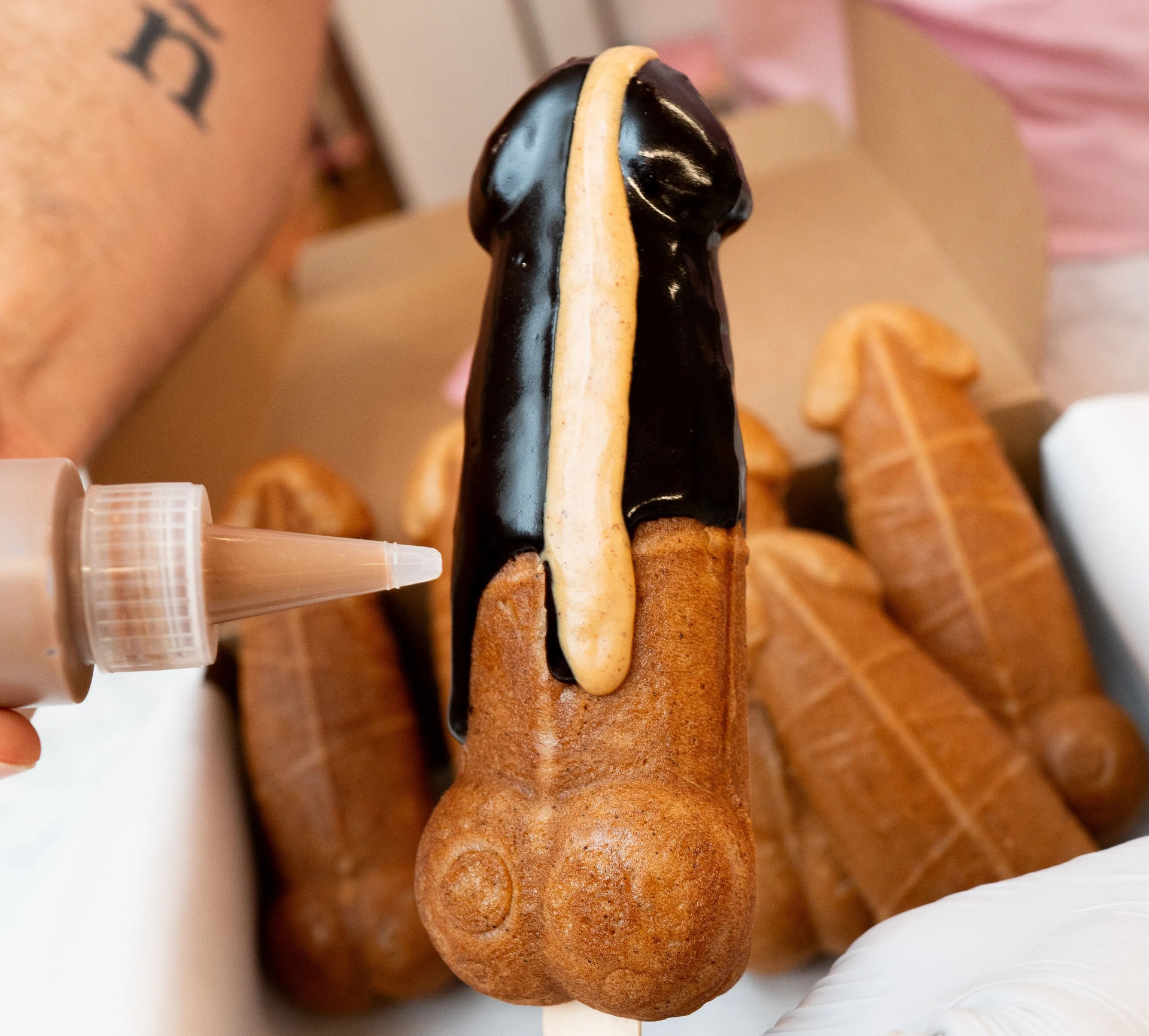 Novelty penis-shaped waffle pop on a stick being drizzled with chocolate and caramel sauce, with a box of similar pastry treats in the background.