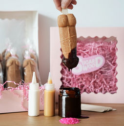 Playful torso-shaped cookie pop on a stick being dipped in glossy chocolate, with squeeze bottles of white and caramel sauce, a chocolate jar, and a pink gift box filled with shredded paper in the background.
