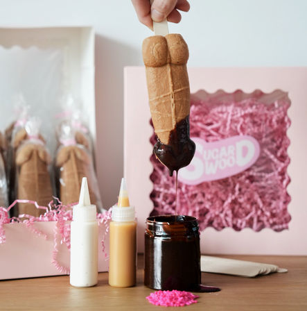 Playful torso-shaped cookie pop on a stick being dipped in glossy chocolate, with squeeze bottles of white and caramel sauce, a chocolate jar, and a pink gift box filled with shredded paper in the background.