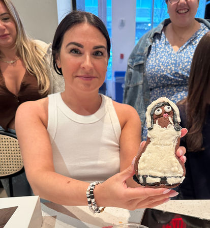 Woman in a white tank top holding a hand-decorated bride-shaped gingerbread cookie with white frosting and candy eyes, friends gathered around a bright indoor counter during a baking get-together.