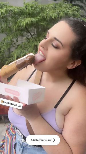 Person in a lilac tank top savoring a chocolate‑dipped ice cream bar on a stick outdoors with a leafy green backdrop