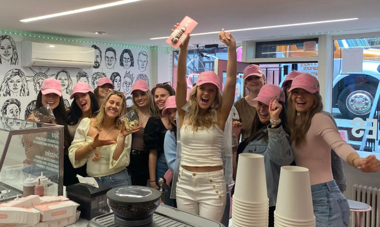 Group of smiling women wearing pink baseball caps celebrating a bachelorette party in a bright ice cream parlor; bride-to-be in white with a veil raises her arms behind a counter with stacked takeaway cups, pastry boxes and a black-and-white mural on the wall.