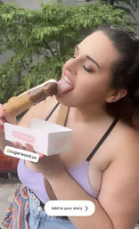 Woman outdoors enjoying a chocolate-dipped ice cream pop on a stick, holding a white dessert box against a leafy background.