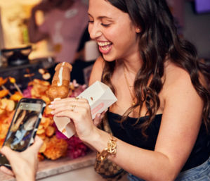 Smiling woman at a party holding a chocolate-drizzled pastry on a stick over a small box as someone photographs it with a smartphone, colorful flowers in the background