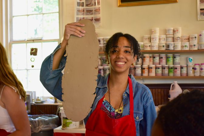 Smiling student in a red apron holding a large slab of clay in a sunlit pottery studio classroom with shelves of glaze jars