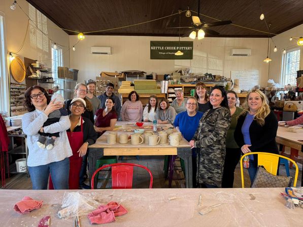 Smiling group at a community pottery class in a bright ceramics studio, gathered around a table of handmade mugs, clay tools, and string lights overhead.