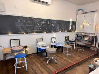 Cozy pottery studio with multiple electric pottery wheels and stools on a worn wooden floor, large chalkboard mural, string lights and shelves of clay supplies