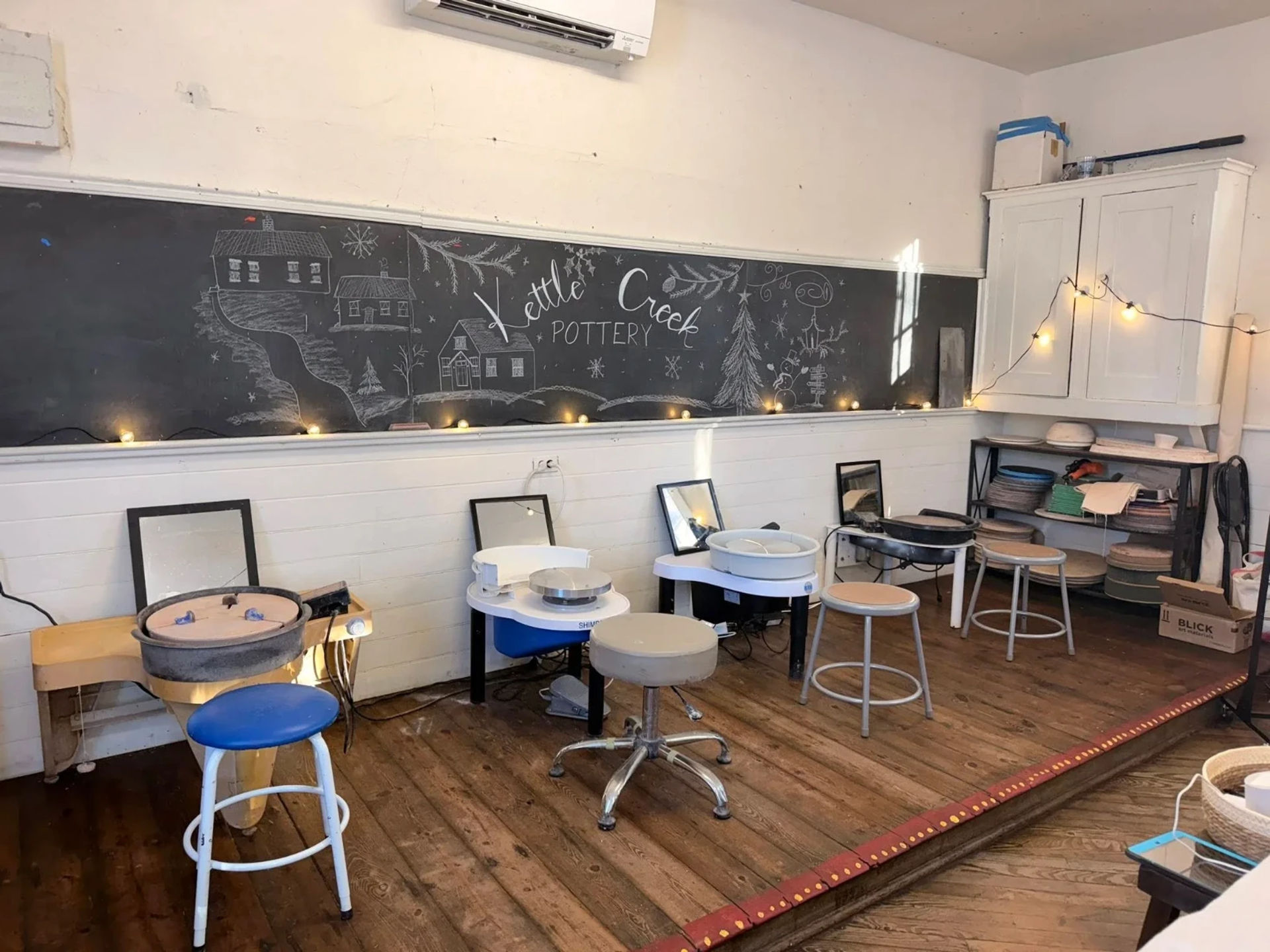 Cozy pottery studio with multiple electric pottery wheels and stools on a worn wooden floor, large chalkboard mural, string lights and shelves of clay supplies