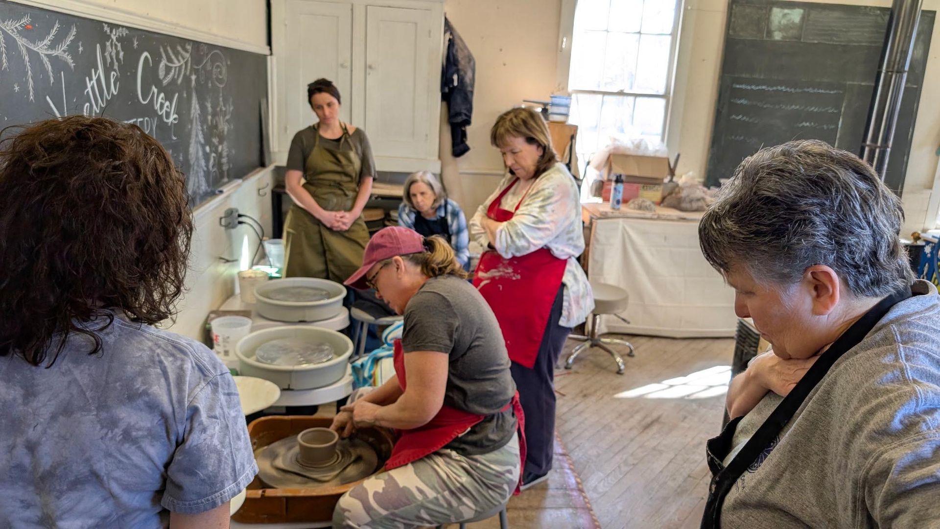 Community pottery class in a sunlit studio: women gathered around a potter shaping clay on a wheel, stacked batts, chalkboard wall and wooden floor visible