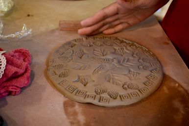 Handmade round clay plaque with pressed leaf, pine cone and butterfly motifs and Greek-key border on a pottery worktable, hand hovering nearby